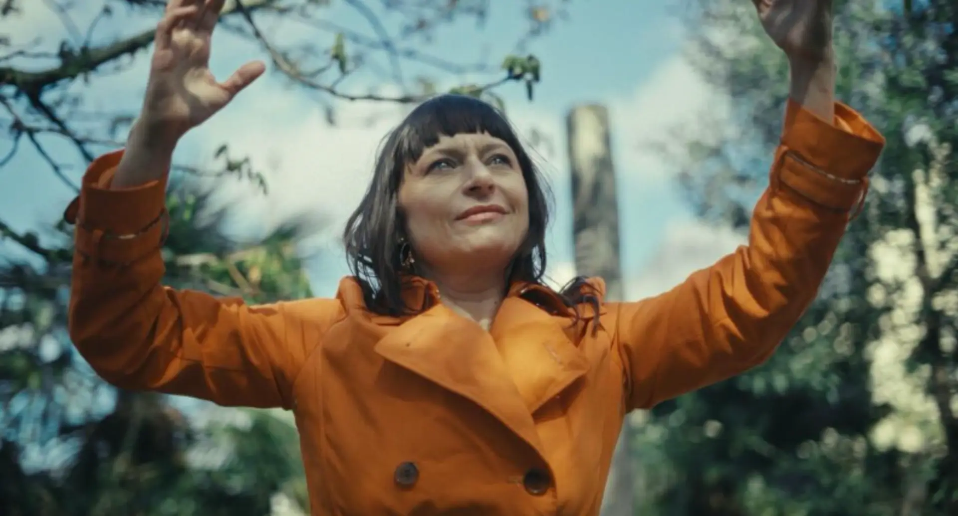 Carole Pellmont dehors devant des arbres avec les bras relevés vers le ciel, elle porte un manteau orange